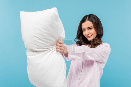 brünette junge Frau im Pyjama hält weißes Kissen isoliert auf blau 
