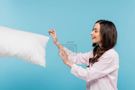 Photo for Side view of happy young woman in sleepwear reaching white pillow in air isolated on blue - Royalty Free Image