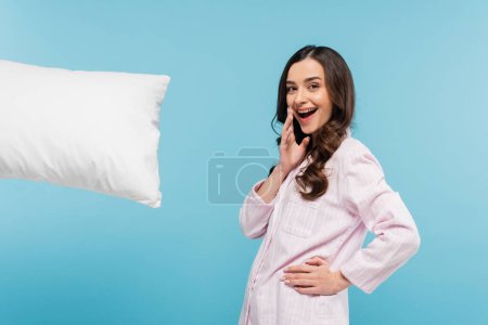 excited young woman in sleepwear standing with hand on hip near levitating pillow isolated on blue 