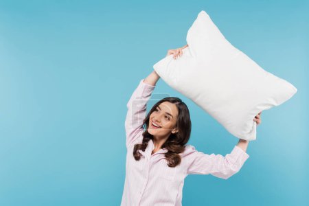 Téléchargez les photos : Joyeuse jeune femme en vêtements de nuit tenant un oreiller blanc au-dessus de la tête isolé sur bleu - en image libre de droit
