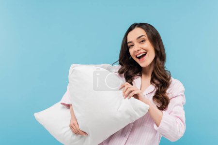 amazed young woman in sleepwear holding soft pillow isolated on blue 