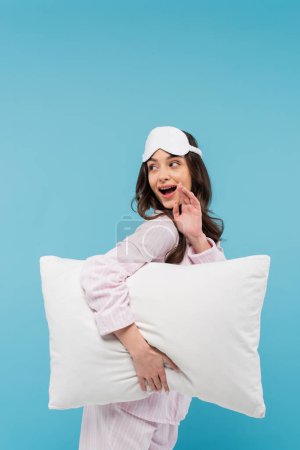 erstaunte junge Frau in Nachtwäsche und Schlafmaske mit weißem Kissen auf blauem Hintergrund 