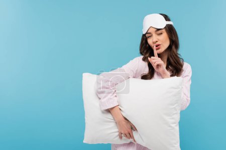 junge Frau in Nachtwäsche und Schlafmaske hält weißes Kissen, während sie Schweigezeichen auf blauem Hintergrund zeigt 