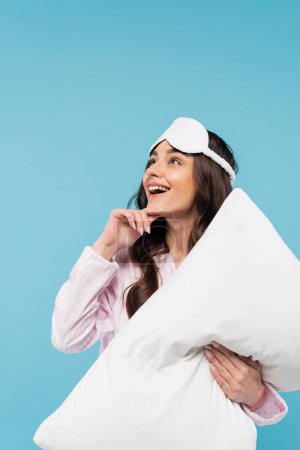 dreamy young woman in nightwear and sleeping mask holding pillow isolated on blue 