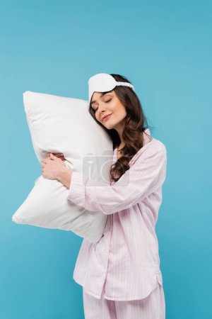 young woman in pajamas and night mask sleeping on white pillow isolated on blue 