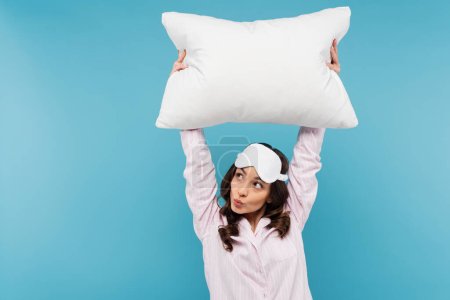 brunette young woman in pajamas and night mask holding white pillow while pouting lips isolated on blue 