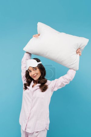 pleased woman in pajamas and night mask holding white pillow above head isolated on blue 