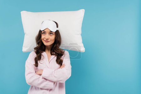 smiling young woman in pajamas and night mask standing with crossed arms near white flying pillow isolated on blue