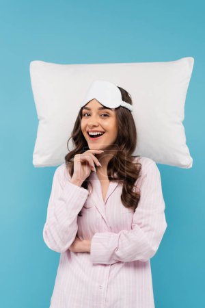 aufgeregte junge Frau in Pyjama und Nachtmaske lächelt neben weißem fliegendem Kissen isoliert auf blauem Grund 