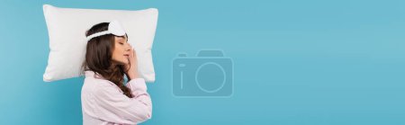 Photo for Side view of brunette woman in pajamas and night mask sleeping on white pillow isolated on blue, banner - Royalty Free Image