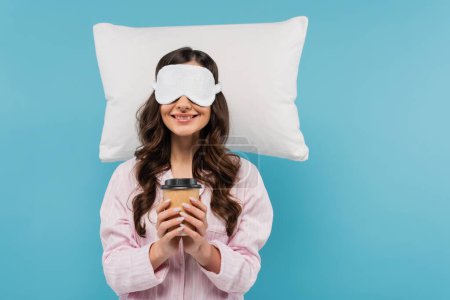 Photo for Pleased woman in pajamas and night mask holding paper cup with coffee to go near white pillow isolated on blue - Royalty Free Image