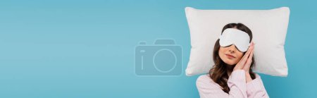 young woman in pajamas and night mask resting on white pillow isolated on blue, banner 