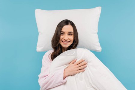 glückliche junge Frau im Pyjama mit warmer Bettdecke neben fliegendem Kissen isoliert auf blauem Grund 