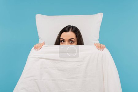 junge Frau im Schlafanzug bedeckt Gesicht mit warmer Bettdecke in der Nähe von Kissen isoliert auf blau 