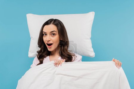 erstaunt junge Frau im Pyjama hält warme Bettdecke neben fliegendem Kissen isoliert auf blau 