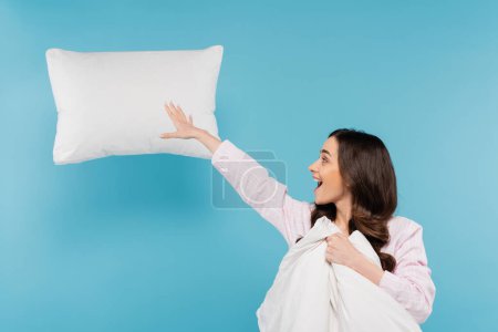 excited woman in pajamas holding warm duvet near flying pillow on blue 