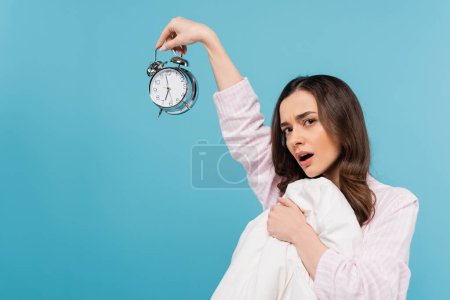shocked young woman in pajamas holding vintage alarm clock and duvet isolated on blue 