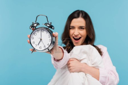 amazed woman in pajamas holding vintage alarm clock and warm duvet isolated on blue 