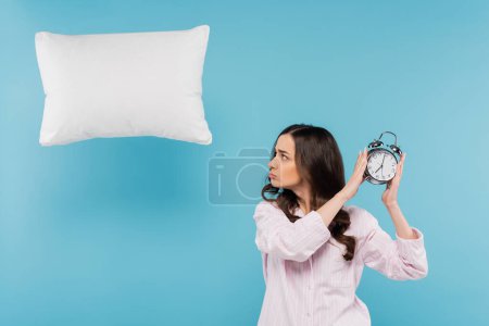 upset young woman in pajamas holding vintage alarm clock near flying pillow on blue 