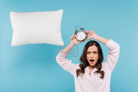 shocked woman in pajamas holding vintage alarm clock above head near levitating pillow on blue 