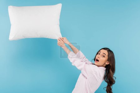 Foto de Shocked woman in pajamas pulling flying pillow on blue background - Imagen libre de derechos