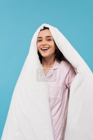 positive junge Frau im Schlafanzug, die den Kopf mit warmer Bettdecke bedeckt und isoliert auf blauem Grund in die Kamera blickt 