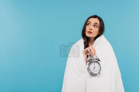 mujer joven soñadora cubierta de edredón blanco sosteniendo reloj despertador vintage aislado en azul 