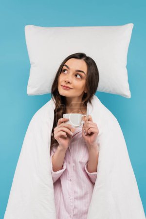 young woman covered in white duvet holding cup near flying pillow isolated on blue 