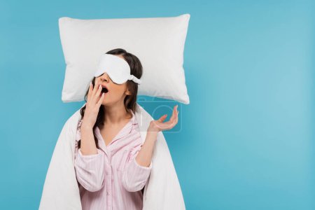 mujer joven con máscara de noche bostezando cerca de almohada levitante aislado en azul 