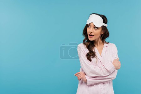 curious young woman in night mask and pajamas looking away isolated on blue 