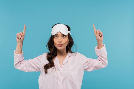 Photo for Brunette young woman in night mask and pajamas looking up and pointing with fingers isolated on blue - Royalty Free Image