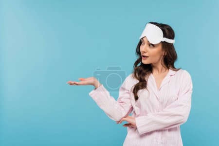 brunette young woman in night mask and pajamas looking away and pointing with hand isolated on blue 