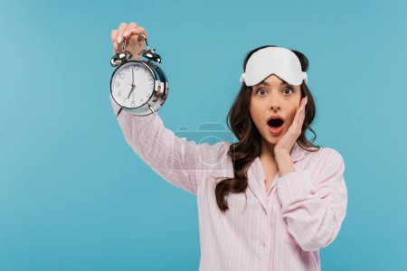 shocked young woman in night mask and pajamas holding alarm clock isolated on blue 