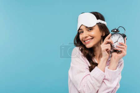 Photo for Pleased young woman in night mask and sleepwear holding alarm clock isolated on blue - Royalty Free Image