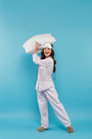Photo for Full length of happy young woman in night mask and sleepwear holding pillow on blue - Royalty Free Image