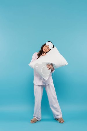 full length of sleepy young woman in night mask and pajamas holding pillow on blue 