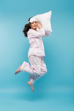full length of barefoot young woman in night mask and sleepwear holding pillow and levitating on blue 