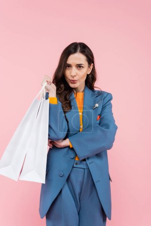 Photo for Young woman in blue blazer holding shopping bags and pouting lips isolated on pink - Royalty Free Image