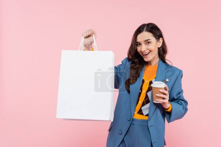 Téléchargez les photos : Heureux jeune femme en blazer bleu tenant des sacs à provisions et café pour aller isolé sur rose - en image libre de droit