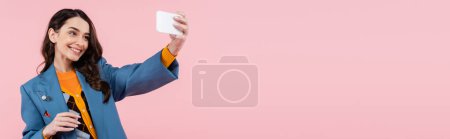 Téléchargez les photos : Happy young woman in blue blazer holding smartphone while taking selfie isolated on pink, banner - en image libre de droit