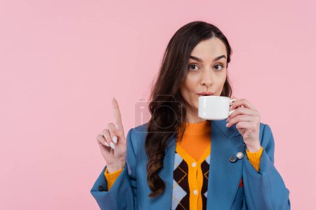 brunette woman in blue blazer drinking coffee and showing wait gesture isolated on pink 