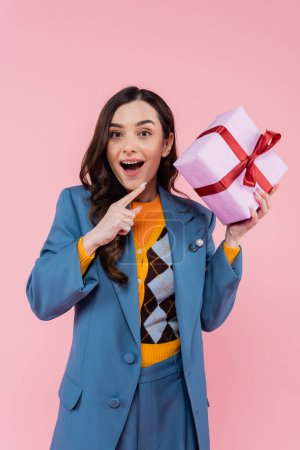 amazed young woman in blue blazer pointing at wrapped present isolated on pink 
