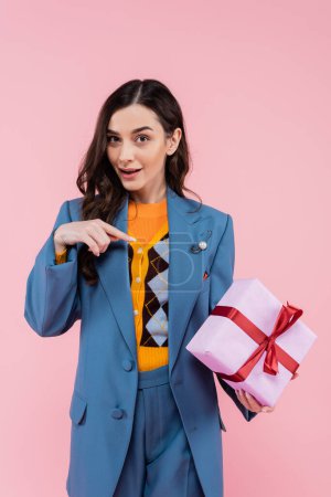 young woman in blue blazer pointing with finger at wrapped present isolated on pink 