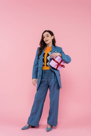 full length of young woman in blue blazer holding wrapped present on pink 