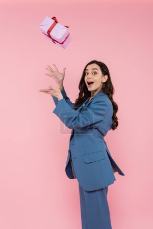 amazed young woman in blue blazer catching wrapped present isolated on pink 