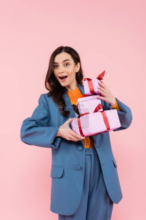 impressed woman in blue jacket holding gift boxes while looking at camera isolated on pink