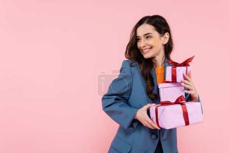 fröhliche brünette Frau im blauen Blazer hält Geschenkboxen in der Hand und schaut isoliert auf rosa