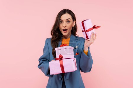 amazed woman in blue stylish blazer holding presents while looking at camera isolated on pink