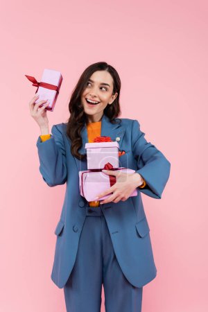 amazed and happy woman in blue suit holding presents while standing with open mouth isolated on pink