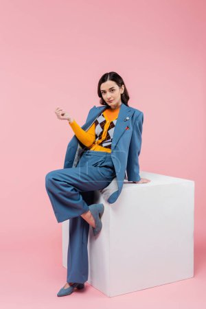 volle Länge der trendigen brünetten Frau im blauen Anzug posiert in der Nähe von White Cube und schaut in die Kamera auf rosa Hintergrund